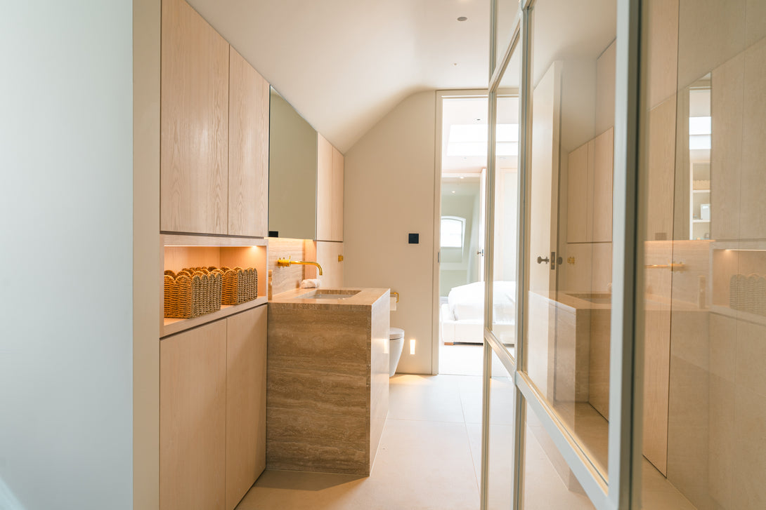 An angled view looking down the long ensuite bathroom, showing the travertine vanity and light oak cabinetry leading to the doorway of the bedroom.