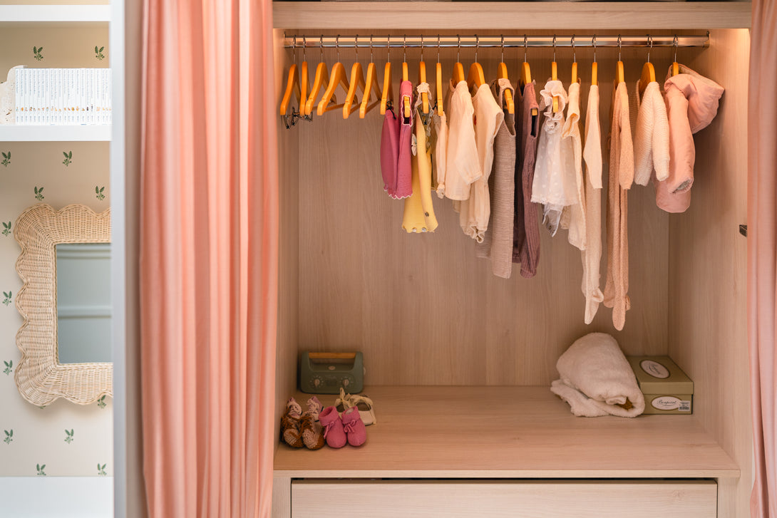 An open view of the baby's bedroom wardrobe, revealing a natural oak interior with a hanging rail full of baby clothes, drawers, and baskets on top, integrated into bespoke cabinetry.