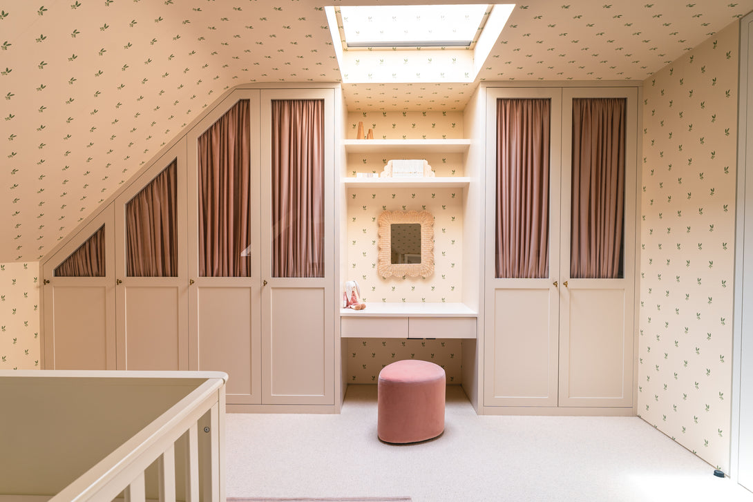A wide view of a baby's bedroom featuring custom-built cream-painted wardrobes, a central dressing table with a woven mirror, and soft pink curtains. The room has sloping ceilings and delicate leaf-patterned wallpaper.