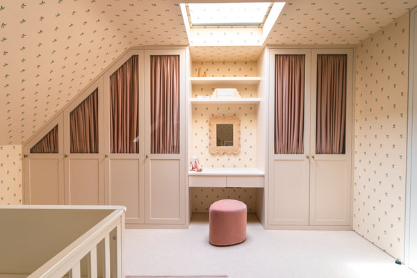 A wide view of a baby's bedroom featuring custom-built cream-painted wardrobes, a central dressing table with a woven mirror, and soft pink curtains. The room has sloping ceilings and delicate leaf-patterned wallpaper.