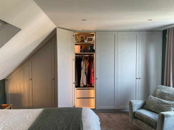 A view from the end of the bed in a loft conversion, showing one of the bespoke wardrobes open to reveal the lit interior with clothes and storage.