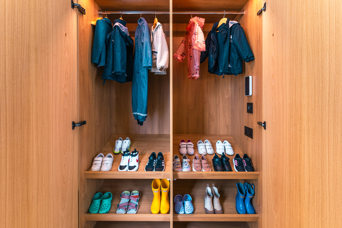 The interior of a bespoke oak cupboard organised for children, with low hanging rails for coats and multiple shelves neatly filled with children's shoes and boots.