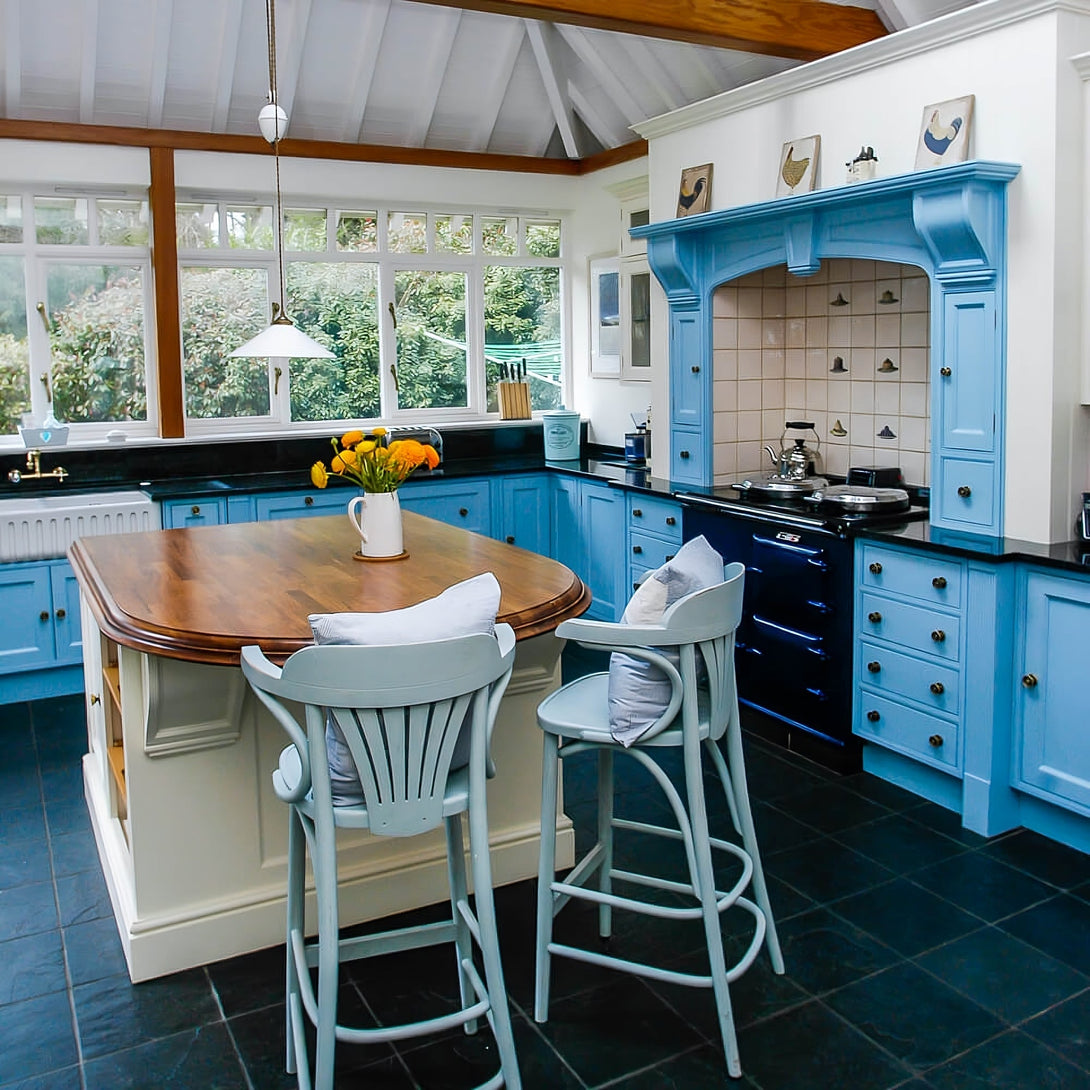 A traditional country-style kitchen featuring hand-painted blue shaker cabinets, a large cream island with a solid wood top, and a bespoke blue mantelpiece over a range cooker.