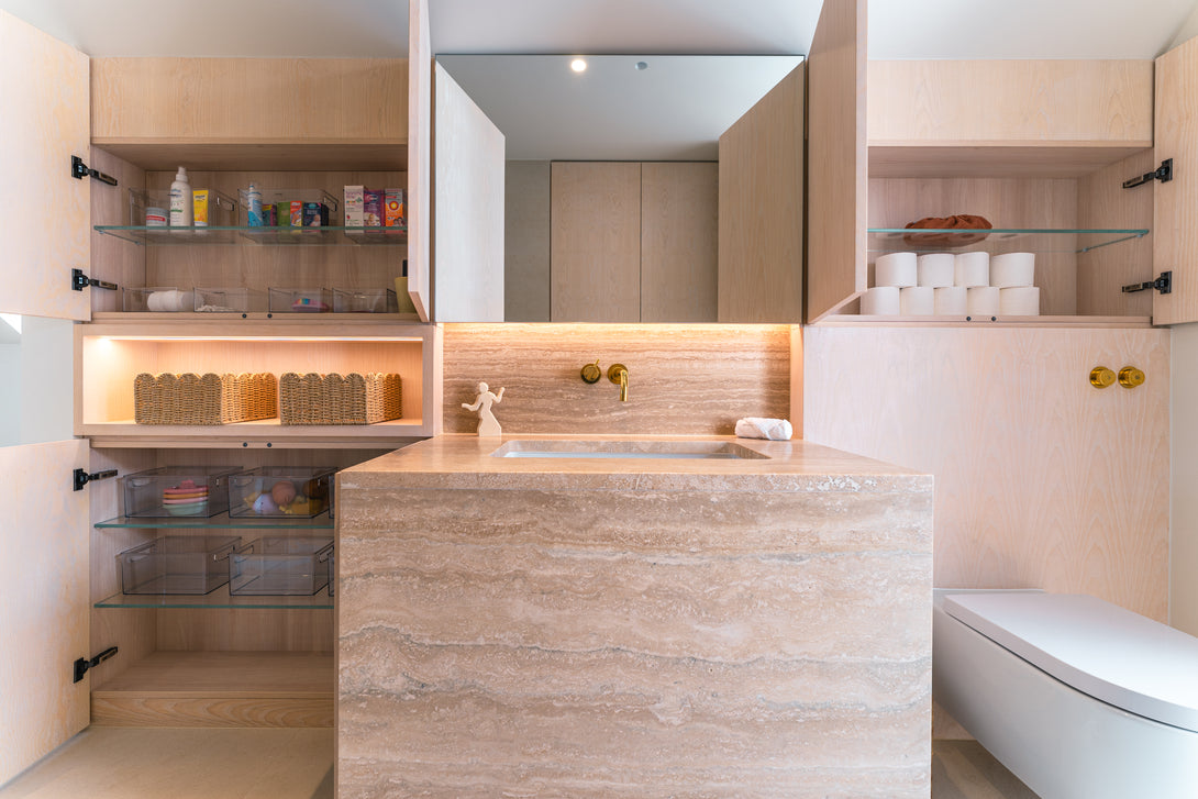 The bespoke light oak bathroom cabinetry is shown with doors open, revealing organised storage on glass shelves for toiletries and toilet rolls.