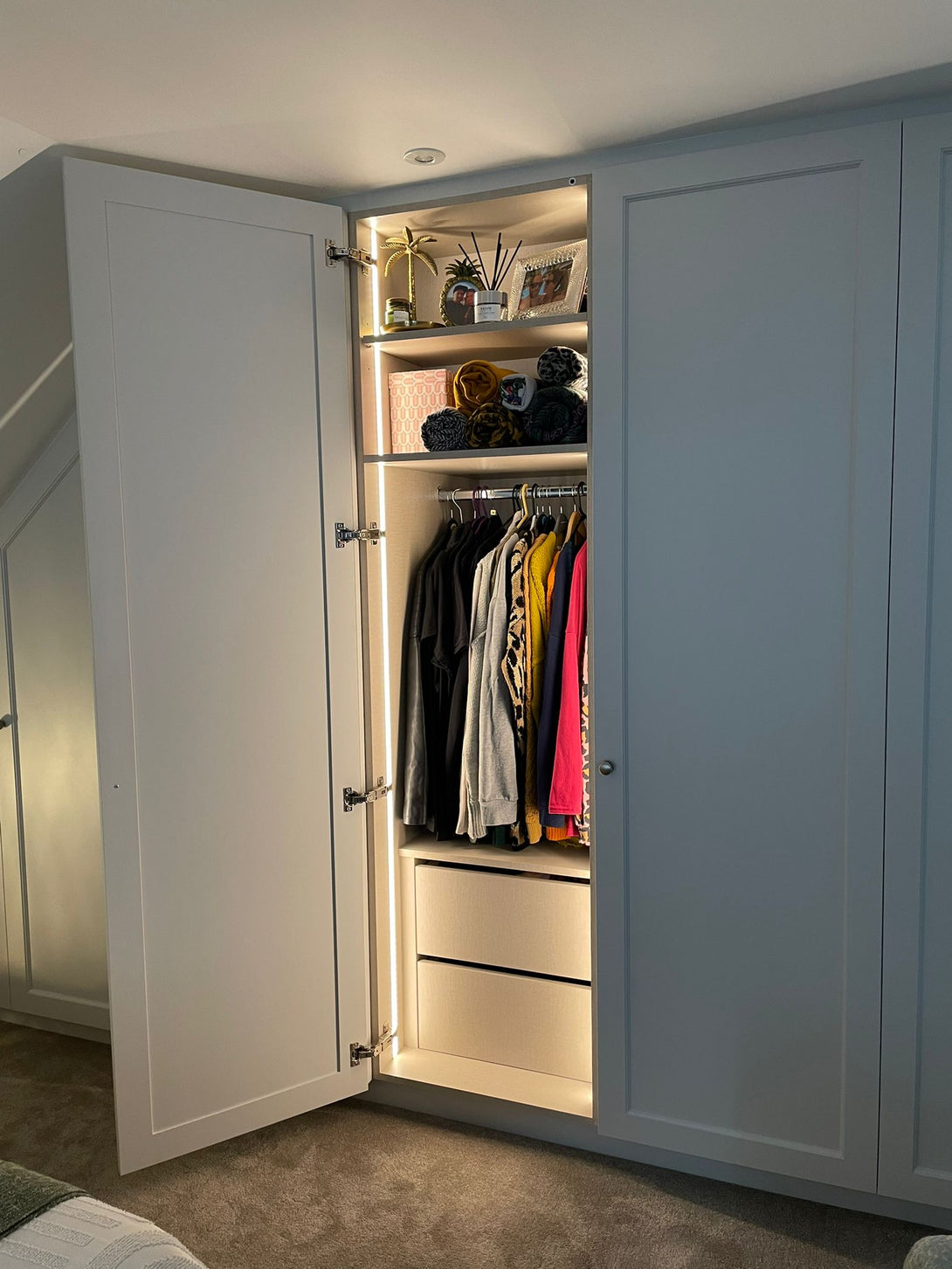 A single bespoke wardrobe open in a loft bedroom, showing the integrated LED lighting illuminating hanging clothes, a display shelf, and drawers.