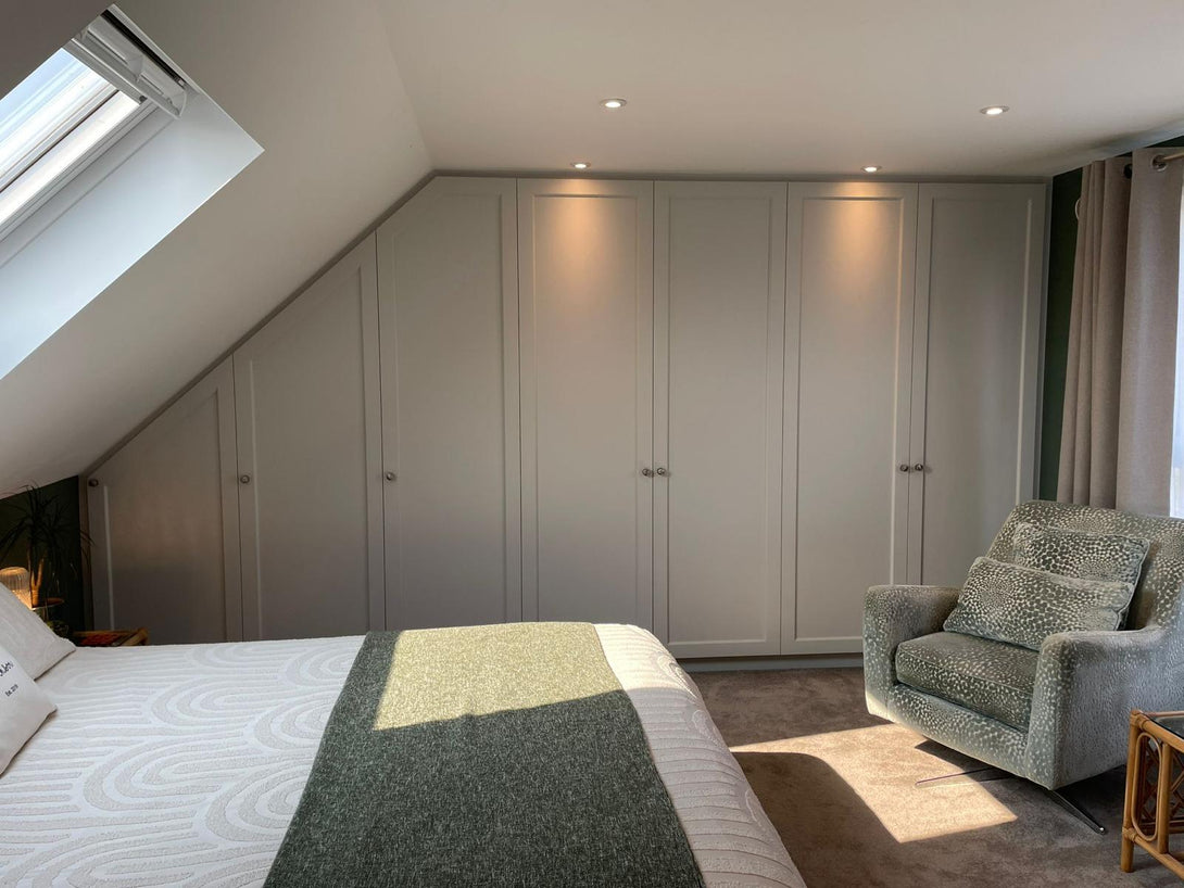 A view of the bespoke angled wardrobes from the bed, with natural light from a skylight highlighting the flawless, soft grey painted finish.