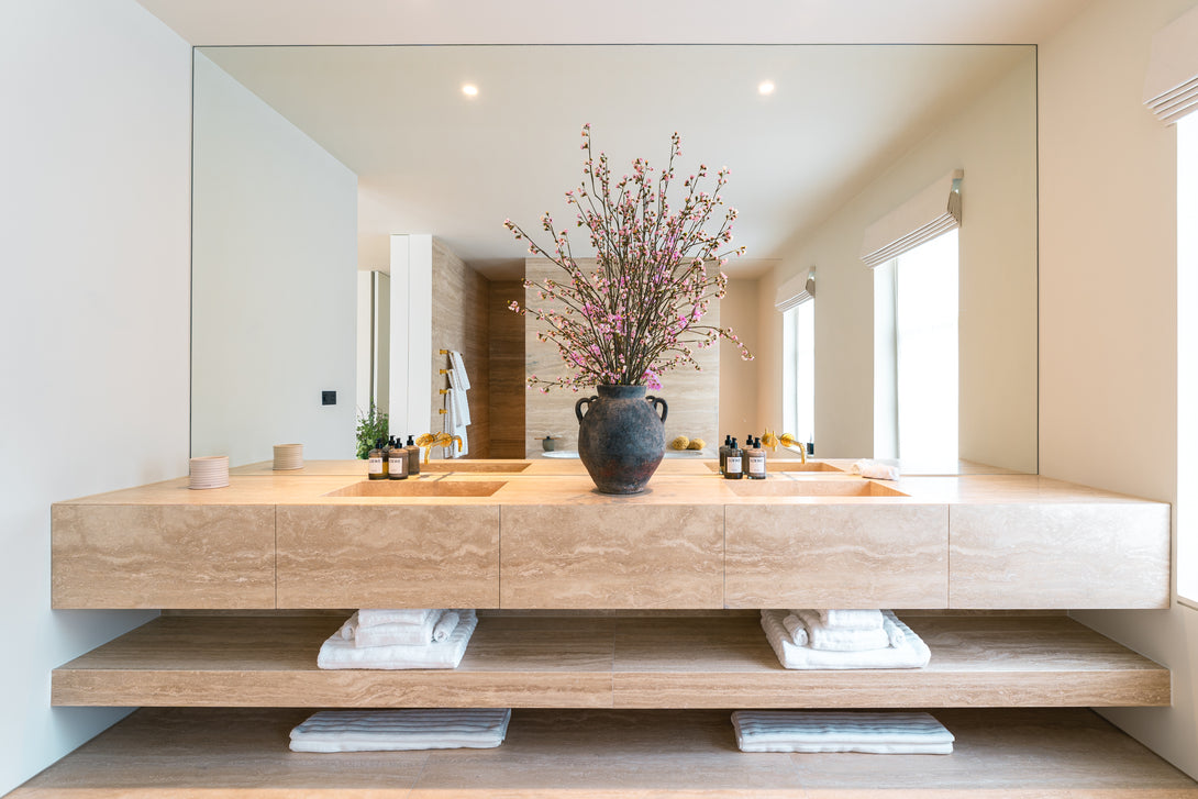 Front view of a bespoke floating travertine double vanity unit in a Master Ensuite by Drew Furniture. The unit features integrated sinks and an open shelf below with folded towels.