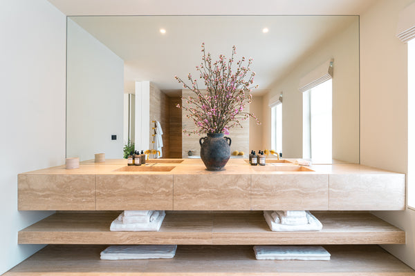 Front view of a bespoke floating travertine double vanity unit in a Master Ensuite by Drew Furniture. The unit features integrated sinks and an open shelf below with folded towels.