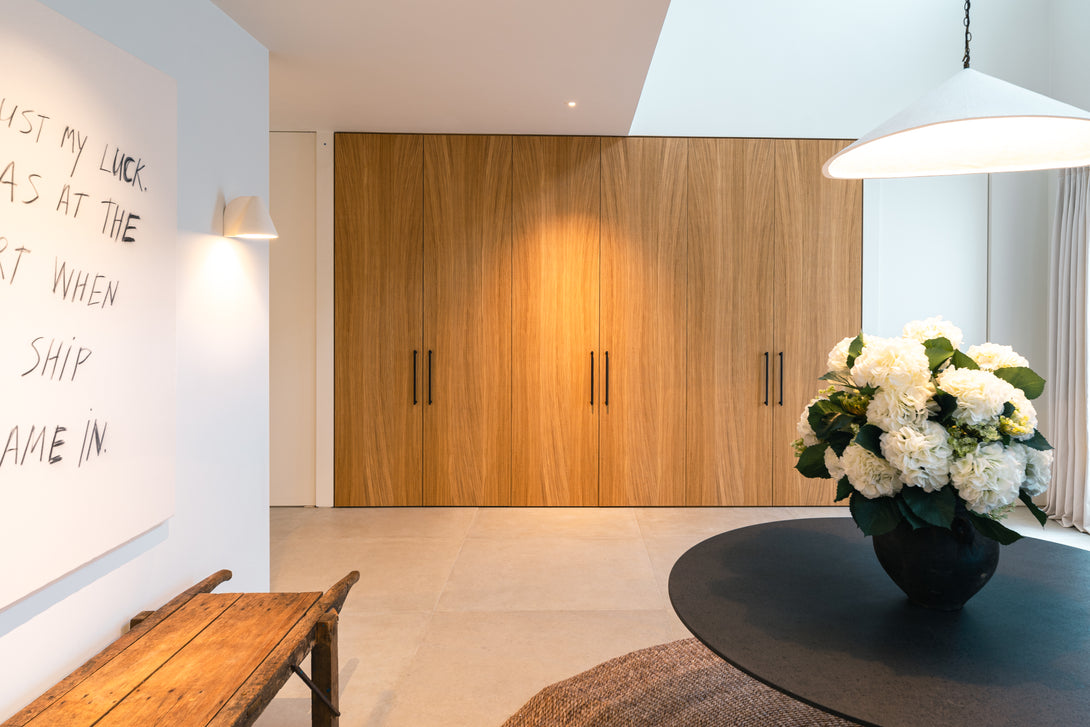 A full wall of bespoke oak cupboards with minimalist black handles shown closed, creating a seamless, architectural wood-panelled look in a modern entrance hall.