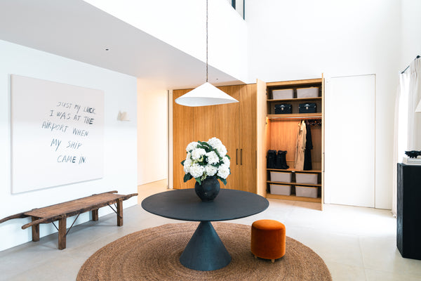 Wide view of a modern, double-height entrance hall featuring a large bank of bespoke oak storage cupboards, a central table with flowers, and contemporary art.