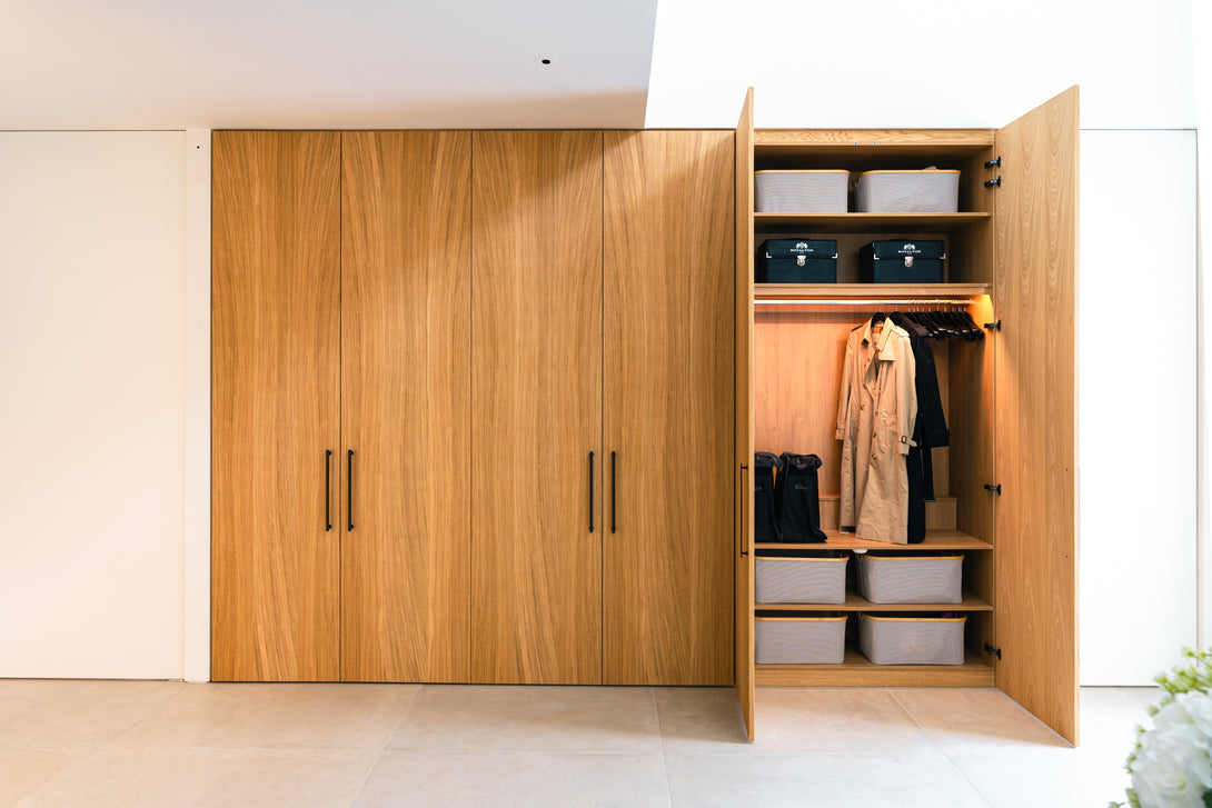 A bespoke floor-to-ceiling oak cupboard in the entrance hall is shown open, revealing integrated lighting, a coat hanging rail, and shelving with storage boxes.