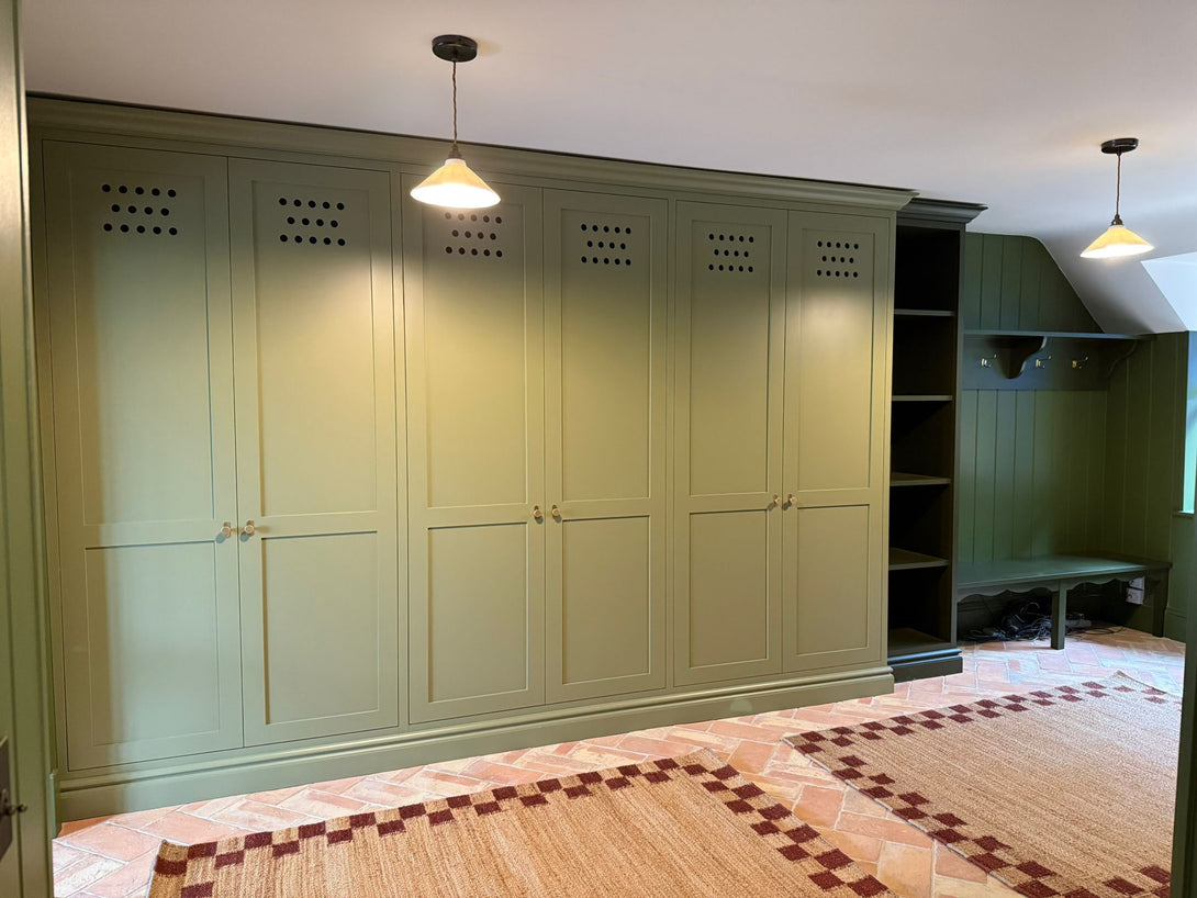 A wide view of the Olive Boot Room by Drew Furniture, featuring a large bank of bespoke, hand-painted olive green storage cabinets and a darker green coat and bench area, set on terracotta flooring.