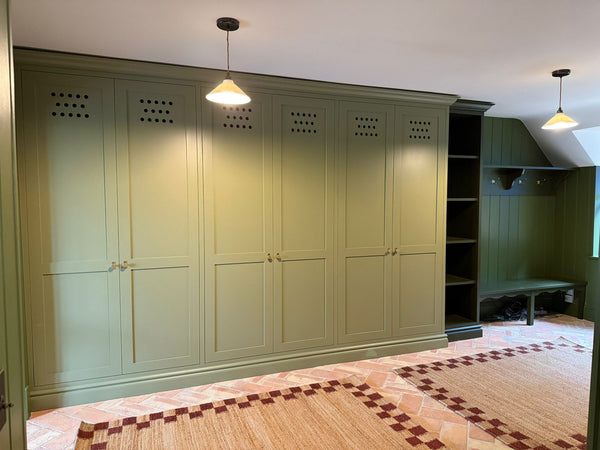A wide view of the Olive Boot Room by Drew Furniture, featuring a large bank of bespoke, hand-painted olive green storage cabinets and a darker green coat and bench area, set on terracotta flooring.