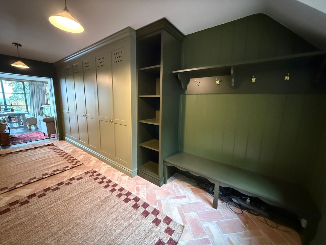 An angled perspective of the bespoke boot room, showing the run of olive green shaker cabinets leading to an integrated dark green bench with coat hooks.