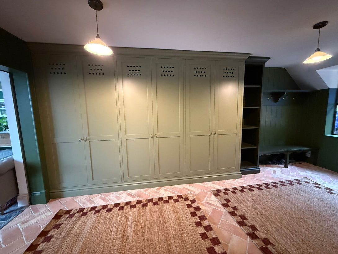 A wide view of the Olive Boot Room by Drew Furniture, featuring a large bank of bespoke, hand-painted olive green storage cabinets and a darker green coat and bench area, set on terracotta flooring.