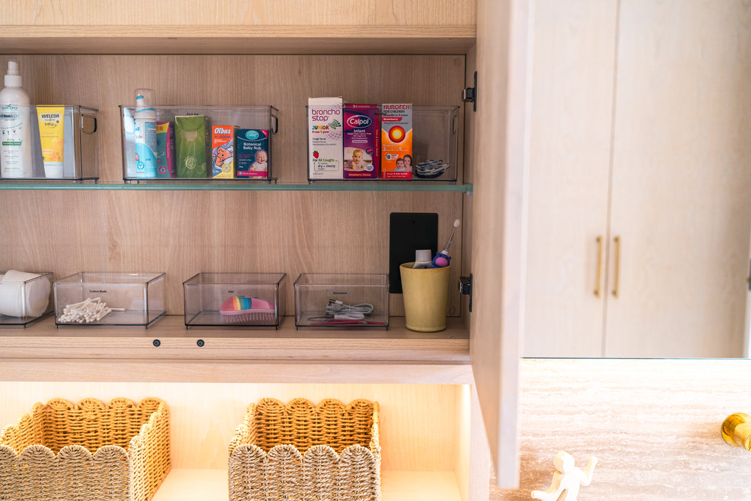 A close-up of an open bathroom cabinet, showing medicines and children's items neatly organised in clear containers on a glass shelf.