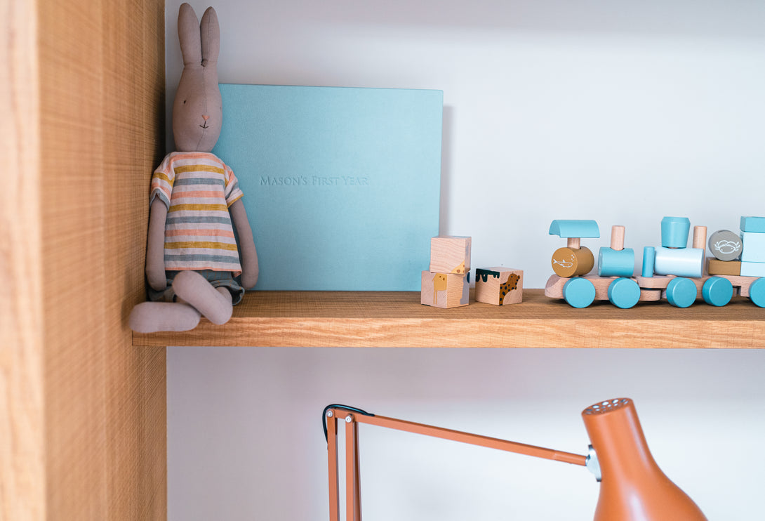 A detailed shot of an oak shelf above the desk, featuring a plush bunny toy, a light blue "Mason's First Year" book, and wooden train toys.