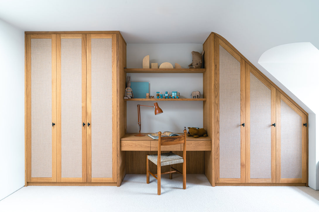 A close-up of a bespoke oak study desk with open shelves displaying toys, flanked by a wardrobe with a woven rattan panel, in a son's bedroom.