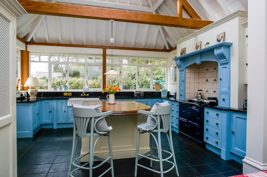 A wide view of a bespoke traditional kitchen with a vaulted ceiling and exposed beams. The shot shows the hand-painted blue cabinets, a cream central island, and large windows.